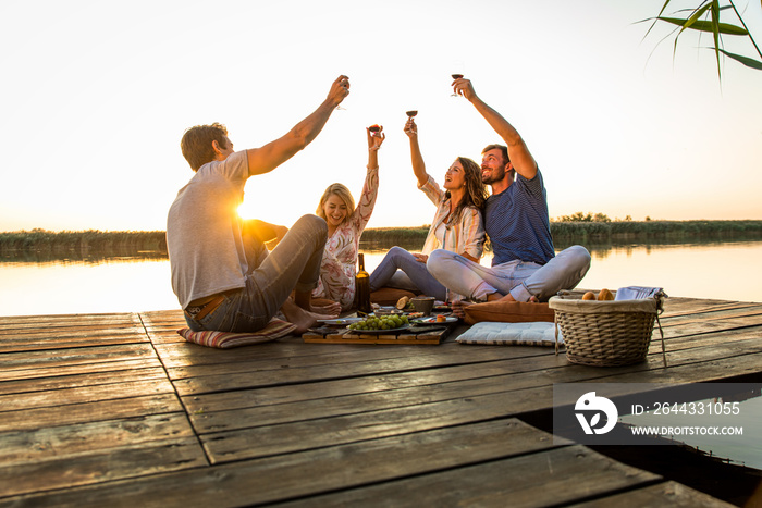 Group of friends having fun on picnic near a lake, sitting on pier eating and drinking wine.