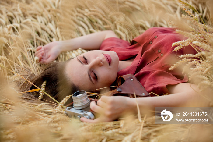 young woman in red dress with camera is lying on spikelets of wheat field