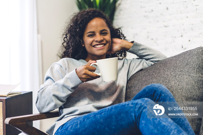 Young african american black woman relaxing drinking cup of hot coffee or tea on couch at home