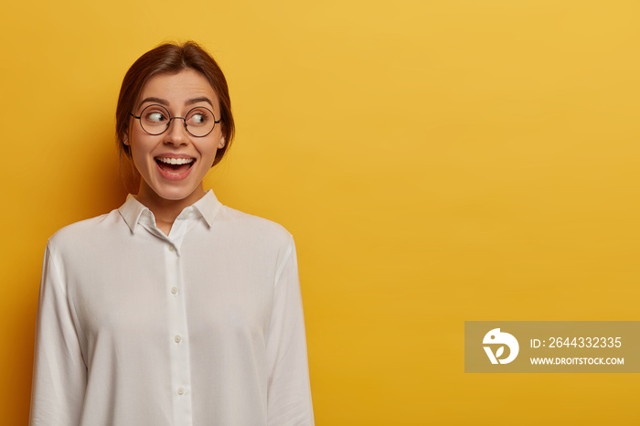 Outgoing pretty female student dressed in white shirt, transparent glasses, happy to finish project work, rejoices after successfully passed exams, looks aside, copy space on yellow background