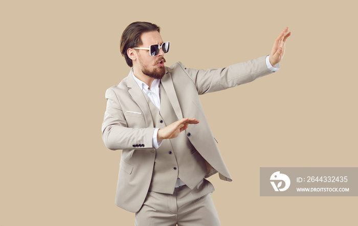 Funny male fashion model in suit having fun in studio. Happy attractive young man wearing modern elegant three piece beige suit and cool sunglasses dancing on beige color background