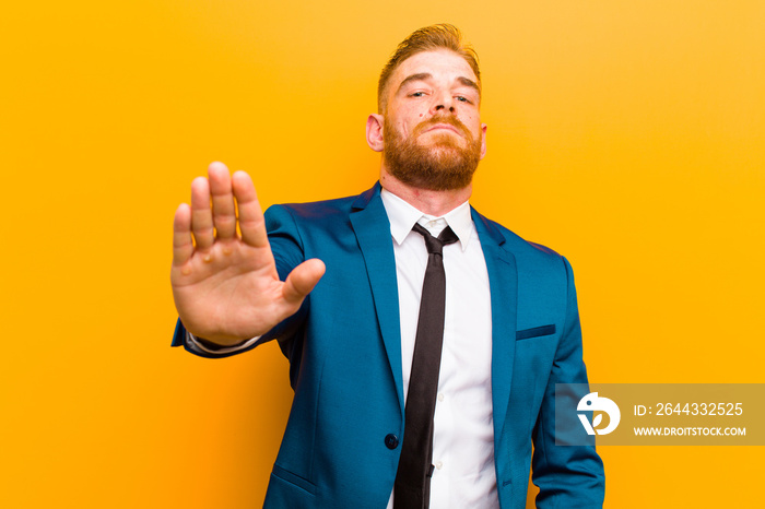 young red head businessman looking serious, stern, displeased and angry showing open palm making stop gesture against orange background