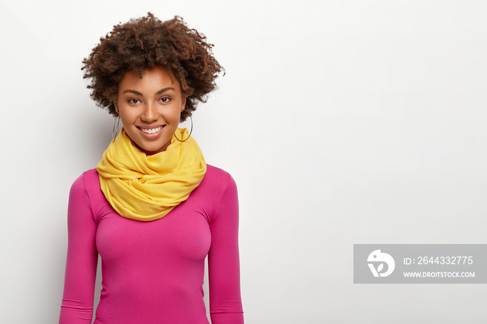 Studio shot of pleasant looking fashionable woman with Afro hairstyle, wears pink sweatshirt and yellow scarf, smiles happily, shows even teeth, being in good mood, gets praise from colleague