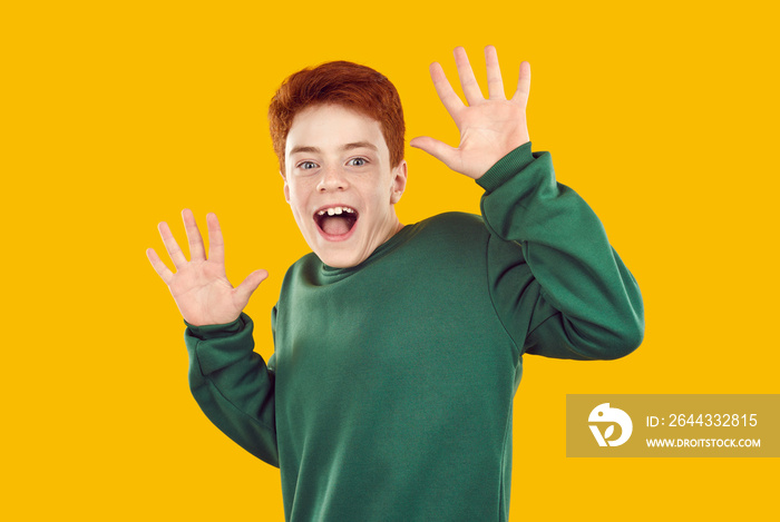 Funny cheerful little redhead child boy in green sweatshirt isolated on amber yellow orange color background sees something unexpected, raises hands and looks at camera with happy surprised expression