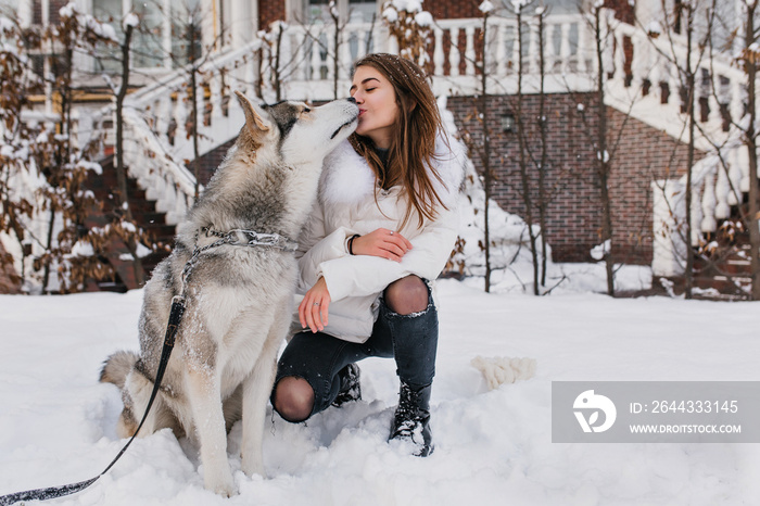 Pretty caucasian lady in white jacket kissing her husky dog during walk in winter day. Attractive young woman wears ripped jeans playing with pet in snowy morning..