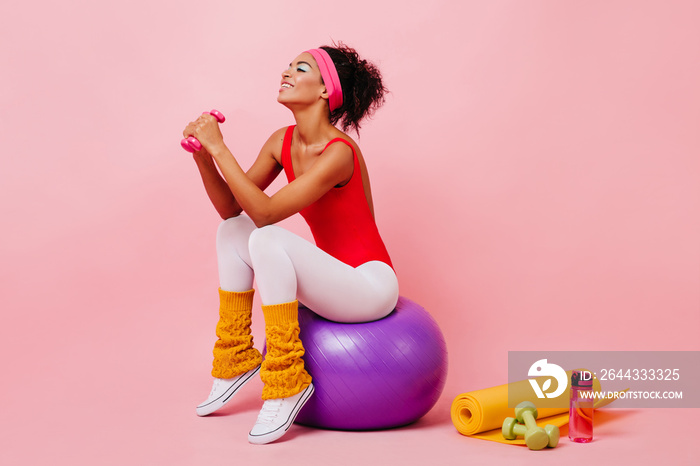 Smiling black girl with dumbbells sitting on pink background. Laughing african sporty woman training with fitness ball.