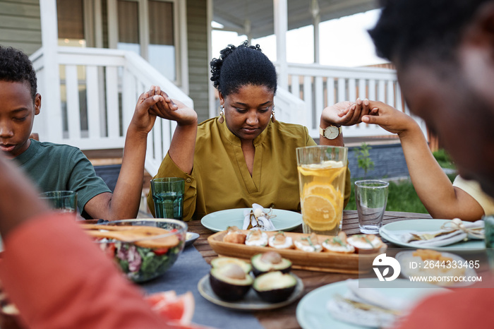 Portrait of young African American woman saying grace at table outdoors during family gathering and holding hands
