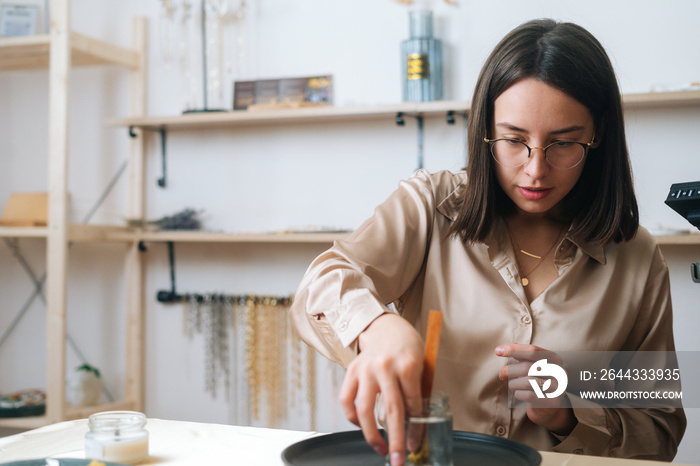 Close-up portrait of focused female artisan preparing glass vessel to pour mixture of liquid wax to create handmade candle in modern workshop. Process of making hand-made natural candle at home.