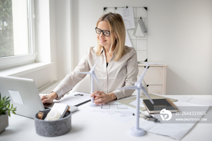 pretty blonde business woman is sitting in her office working on new alternative energy development looking at a model of a wind turbine and is happy and motivated