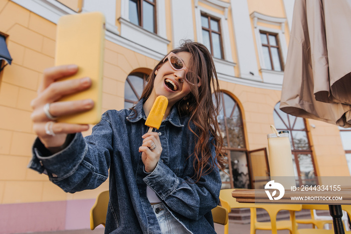 Trendy girl with dark hair in sunglasses eats ice cream on street. Cheerful teenager makes photo on modern gadget, dressed in denim jacket. Youth style, leisure. Concept communication by phone