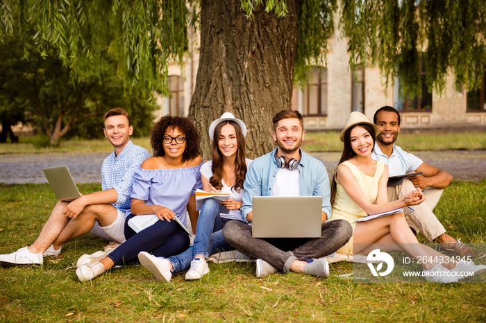 It`s more easy together! Six cheerful classmates are sitting on plaid on the green grass in spring park near campus and study, preparing for exams, tests, looking in camera and smiling