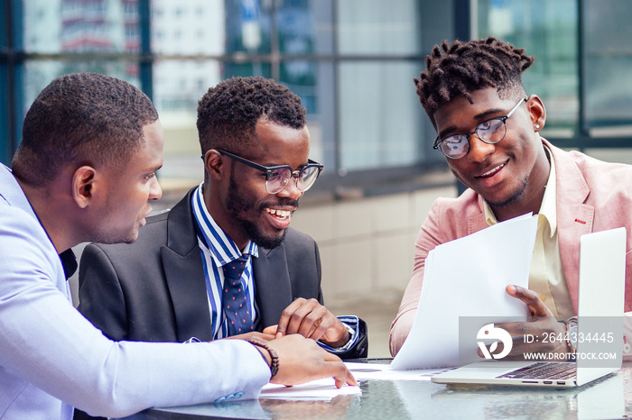 A group of three stylish African American businessman partners entrepreneurs in fashion business suits meeting sitting at table sign securities with laptop in a summer cafe outdoors