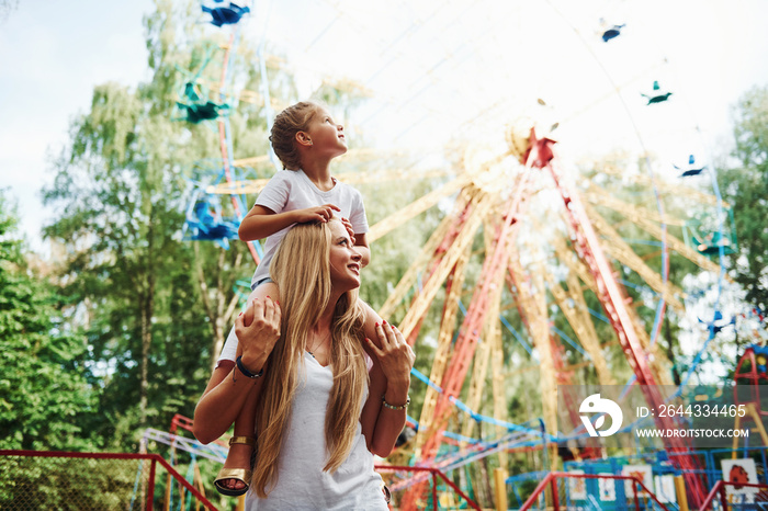 Daughter sits on the shoulders. Cheerful little girl her mother have a good time in the park together near attractions