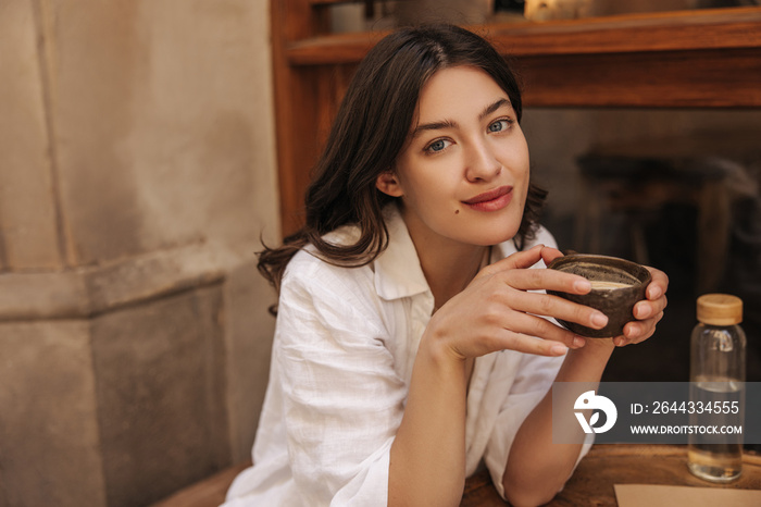 Pretty young caucasian woman looking at camera enjoying coffee in street cafe. Brunette with wavy hair wears white shirt. Lifestyle concept