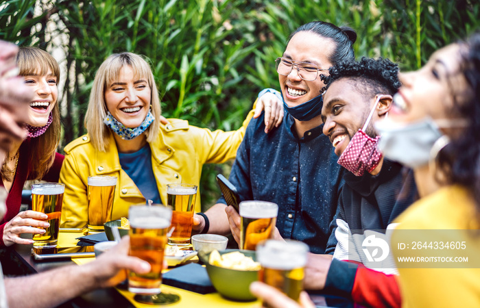 Young people drinking beer with opened face mask - New normal life style concept with millenial friends having fun together on happy hour at brewery garden party - Vivid filter with focus on asian guy