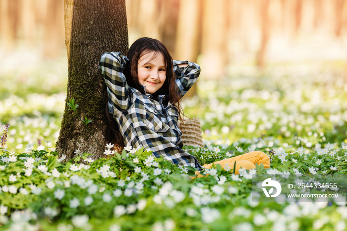 Little happy girl among spring flowers in forest or park. Child is resting under tree.