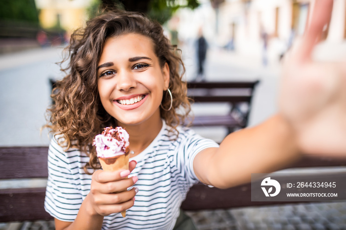 Portrait of young woman eating ice cream and taking selfie photo on camera in summer street.