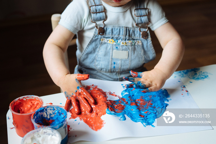 Child painting with her hands on the table at home using blue and red paint. Finger painting or art therapy for children. Fun activities for toddlers. Close up.