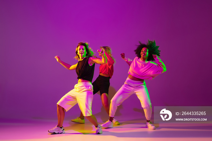 Flexible. Sportive girls dancing hip-hop in stylish clothes on purple-pink background at dance hall in green neon light. Youth culture, movement, style and fashion, action. Fashionable portrait.