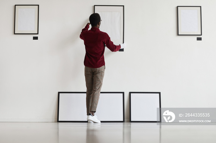 Full length back view at African-American man hanging blank frames on wall while planning art gallery or exhibition, copy space