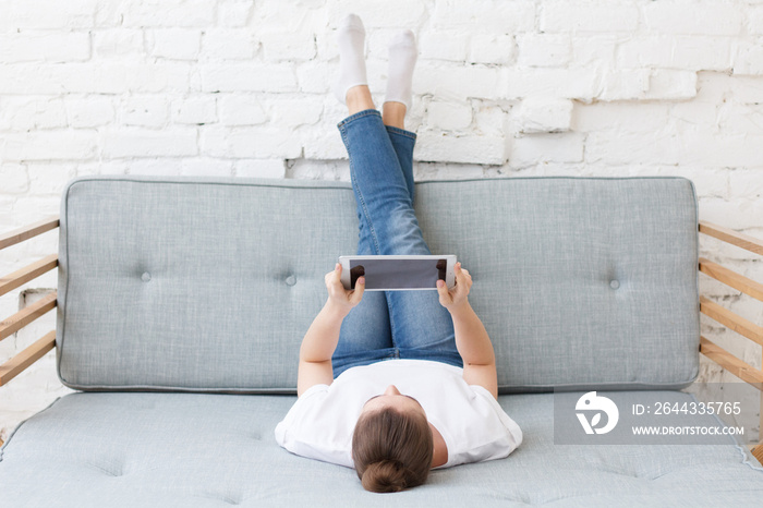 Young woman laying on couch in loft interior, watching movie or playing video game with her tablet pc