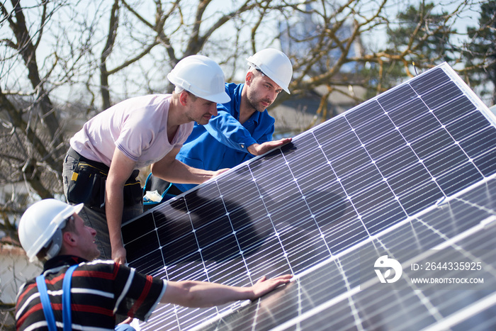 Male team workers installing solar photovoltaic panel system. Three electricians mounting blue solar module on roof of modern house. Alternative energy ecological concept.