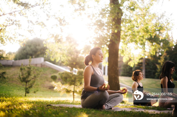 Young woman is meditating in morning in park while sunrise. Group of people outdoors is sitting in lotus pose on green grass with their eyes closed and meditating