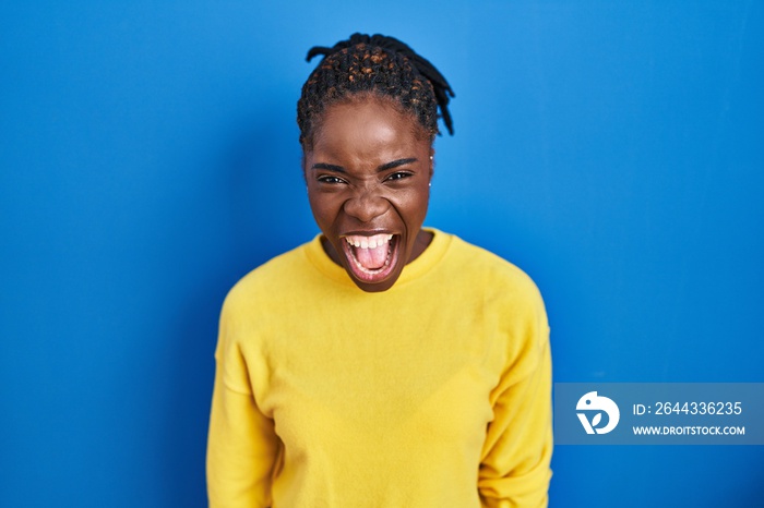 Beautiful black woman standing over blue background angry and mad screaming frustrated and furious, shouting with anger. rage and aggressive concept.