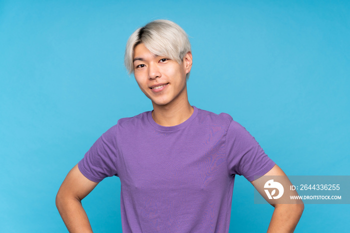 Young asian man over isolated blue background posing with arms at hip and smiling