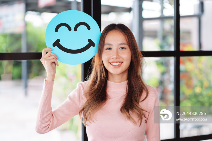 Asian woman holding blue happy smile face on paper cut, user giving good feedback rating, think positive , customer review, assessment, of mental health day, Compliment Day, satisfaction concept.