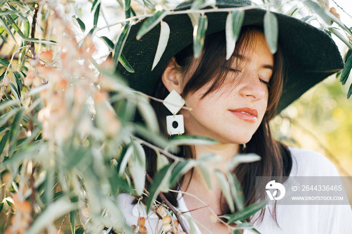 Stylish boho girl in hat and with modern earrings posing among green olive branches in soft evening light. Young fashionable woman relaxing on tropical island. Summer vacation. Space for text