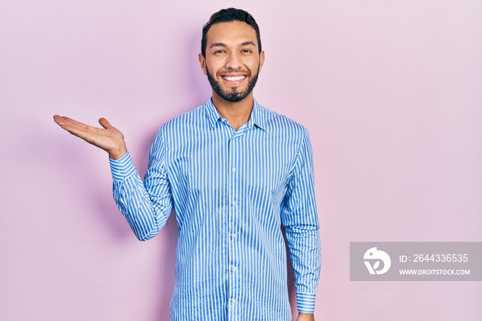 Hispanic man with beard wearing casual blue shirt smiling cheerful presenting and pointing with palm of hand looking at the camera.