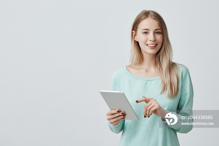 Portrait of attractive young woman with dark shining eyes and blonde long hair wearing light blue sweater working on tablet. Young female entrepreneur with tablet smiling at camera