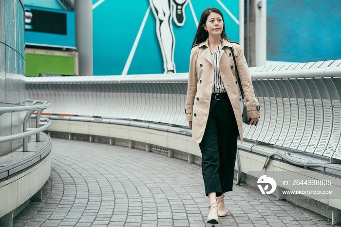 full length of confident asian girl worker walking on bridge at sunset going home from work. young japanese woman crossing overpass wearing high heels shoes. smiling female carrying small bag.