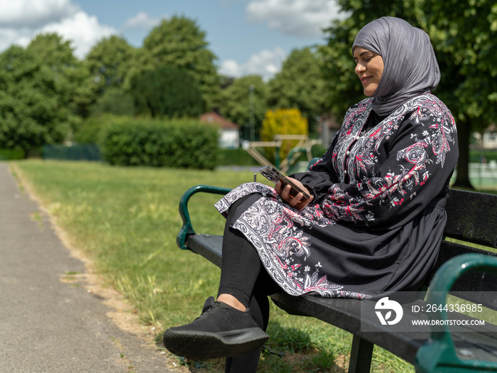 UK,Sutton,Woman in headscarf sitting on bench in park,looking at smart phone