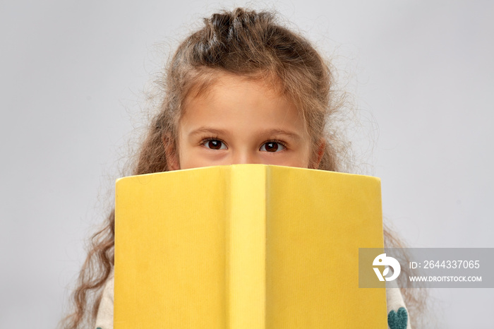 reading, education and childhood concept - little girl hiding behind yellow book over grey background