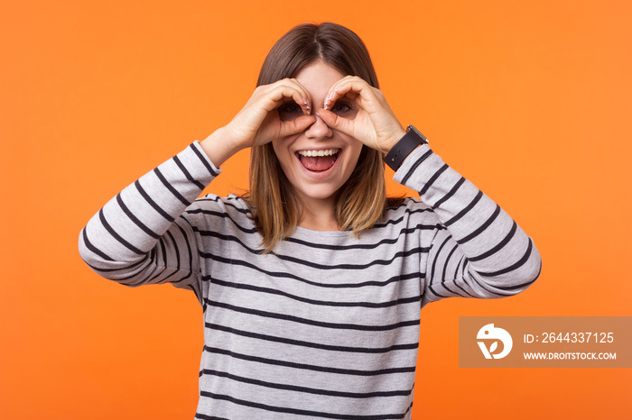 Portrait of curious positive woman with brown hair in long sleeve striped shirt standing, holding hands near eyes like spectacles, binocular gesture. indoor studio shot isolated on orange background