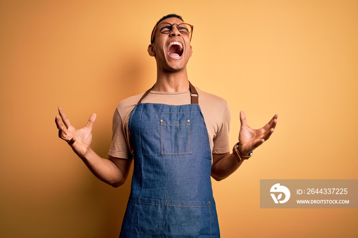 Young handsome african american shopkeeper man wearing apron over yellow background crazy and mad shouting and yelling with aggressive expression and arms raised. Frustration concept.