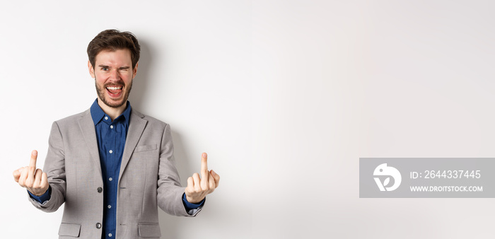 Rude ignorant guy in business suit showing middle fingers and tongue, smiling while mocking people, fuck you gesture, standing on white background