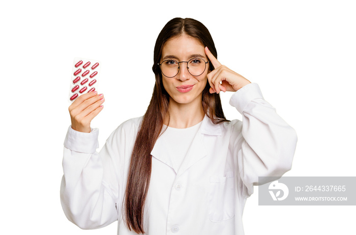 Young pharmacist woman holding a tablet of pills isolated pointing temple with finger, thinking, focused on a task.