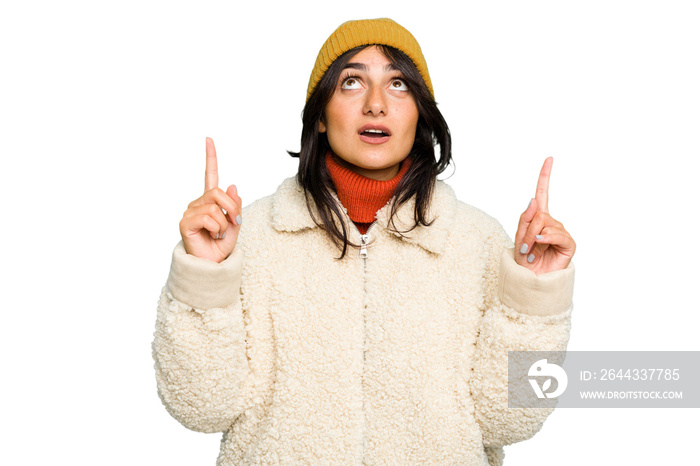 Young Indian woman wearing winter jacket and a wool cap isolated pointing upside with opened mouth.