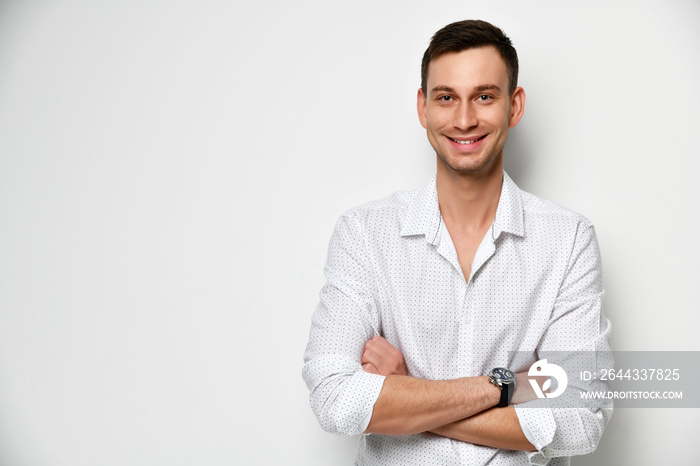 Young man with a wide kind smile stands at the white wall with arms crossed near free text space