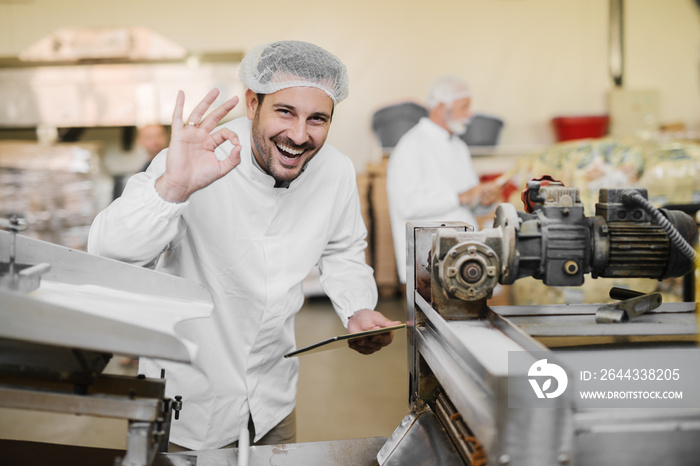 Good quality. Picture of cheerful smiling young man in sterile clothes in food factory. Holding tablet in one hand and with other gesturing that the quality of product is good.