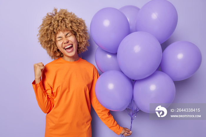 People special occasion and celebration concept. Optimistic woman wears orange jumper clenches fist celebrates special occasion holds bunch of inflated balloons isolated over purple background.