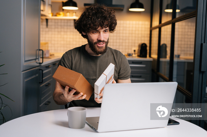 Portrait of happy surprised young curly man opening gift box with present during video call on laptop computer sitting at table in kitchen with modern interior. Concept of remote celebration.