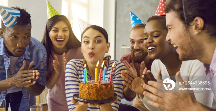 Young people having fun and celebrating friend’s birthday. Banner with group of multiethnic friends applaud as birthday girl in her 20s twenties makes funny face and blows candles lit on party cake