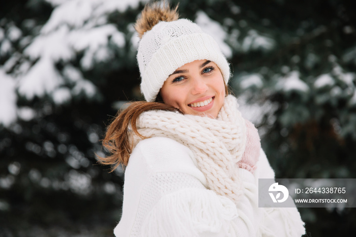 Outdoor close up portrait of young beautiful happy smiling girl wearing white knitted beanie hat, scarf and gloves. Model posing in park with Christmas lights. Winter holidays concept.