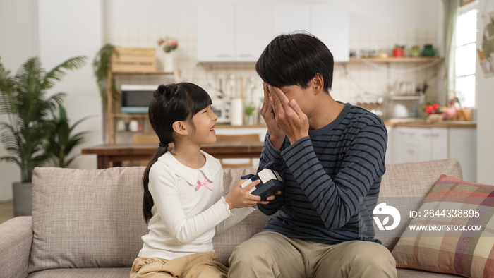 cute asian girl telling dad to cover face and giving him a surprise gift on Father’s Day. they sit on living room sofa together in bright home interior