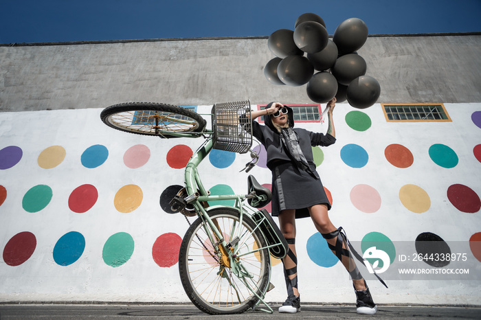 Beautiful young woman jumping with a lot of black baloons and Guy with bicycle posing on the street and looking at she.