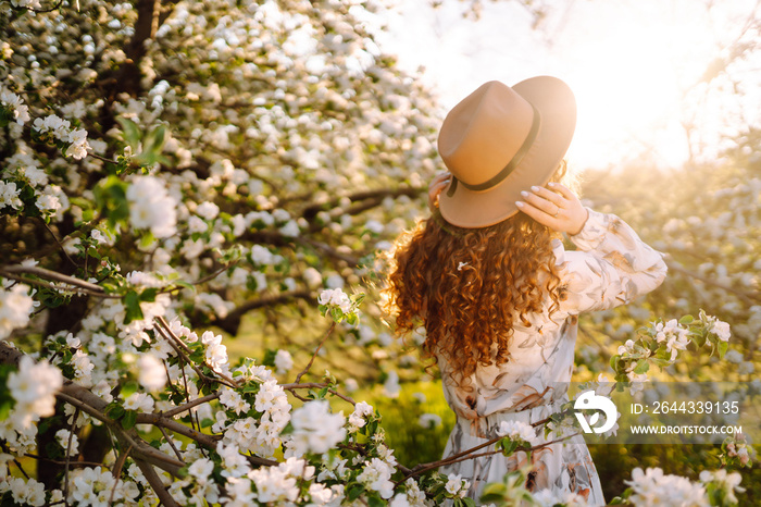 Curly woman with the hat posing in blooming spring park. The concept of relax, travel, paradise, romance and spring vacation. Fashion style.